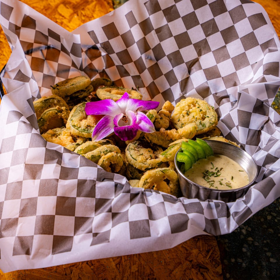 Crispy fried vegetable fritters served with creamy dipping sauce, sliced avocado, and a purple orchid on checkered paper.
