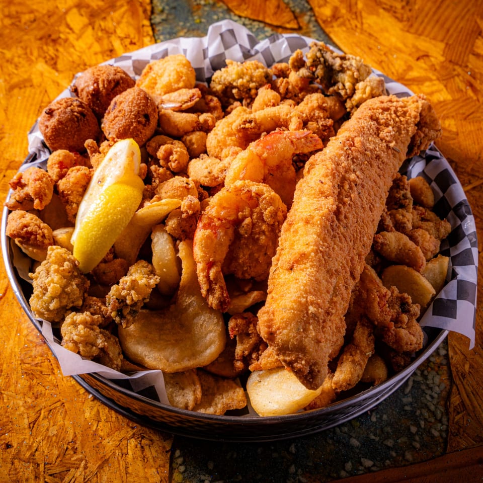 Basket of assorted fried seafood, including fish, shrimp, oysters, and calamari, with lemon and fries on a wooden table.