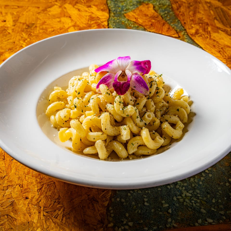 Creamy herb-coated cavatappi pasta served on a white plate, garnished with a purple orchid, atop a rustic wooden table.