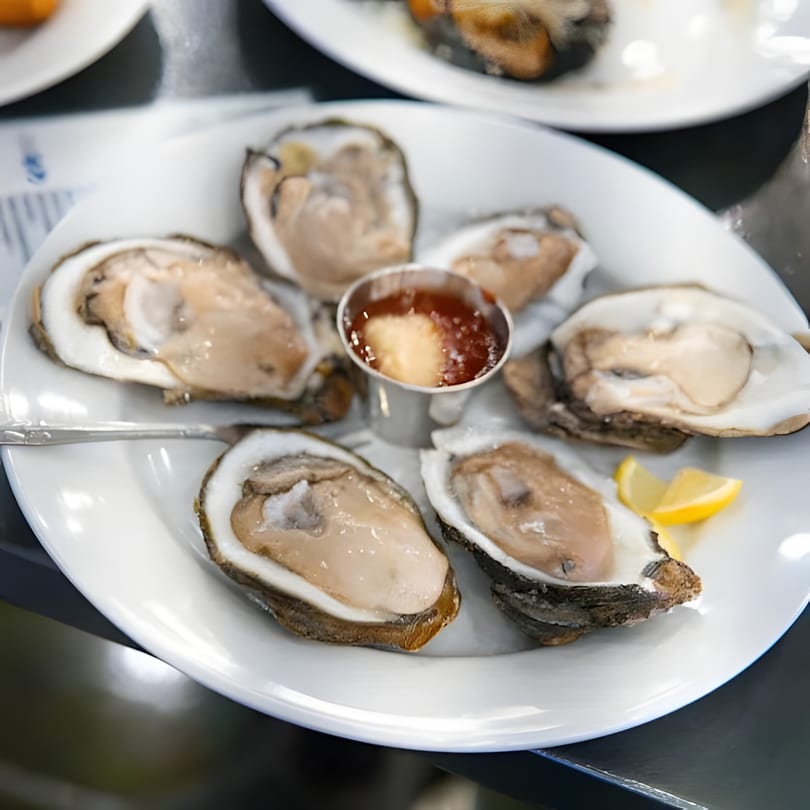 A white plate holds six fresh oysters on the half shell with cocktail sauce and lemon wedges.