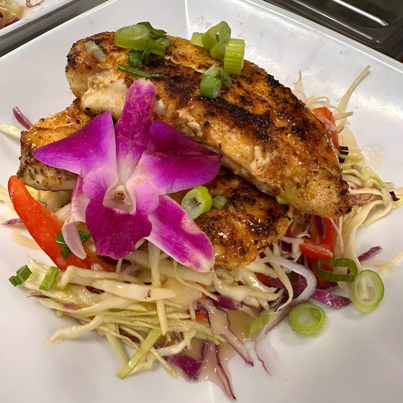 Grilled fish fillets with green onions and a purple orchid, served over a colorful slaw.