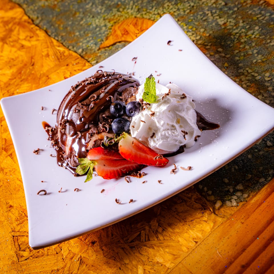 Chocolate dessert topped with chocolate sauce, whipped cream, blueberries, strawberries, mint, and chocolate shavings on a white plate.