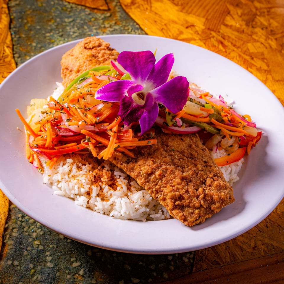 Breaded fish fillet served with white rice, colorful shredded vegetables, and an orchid garnish on a plate.