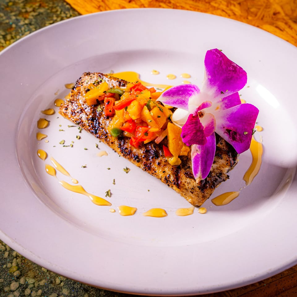 Grilled fish fillet topped with colorful sautéed vegetables, purple edible flowers, and drizzled sauce on a white plate.